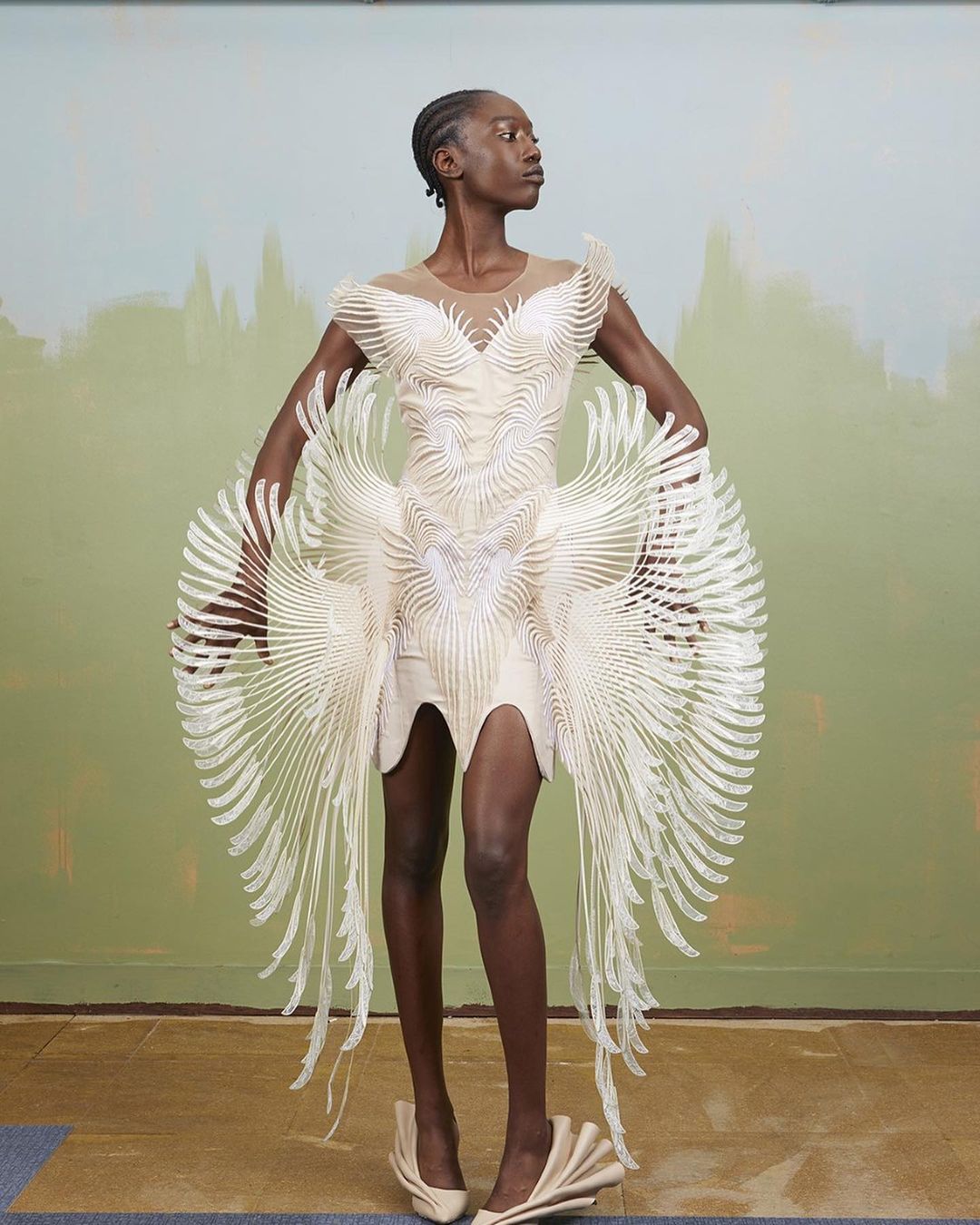 Fashion model poses wearing a sculptural cream dress with flowing accents, paired with matching shoes against a painterly backdrop.