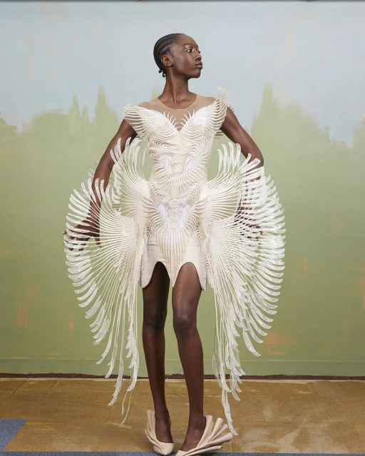 Fashion model poses wearing a sculptural cream dress with flowing accents, paired with matching shoes against a painterly backdrop.