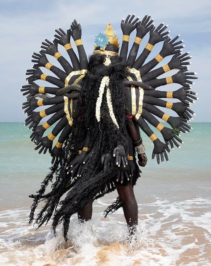 Person in black body paint and costume with multiple arms stands on a beach with ocean and sky in the background.