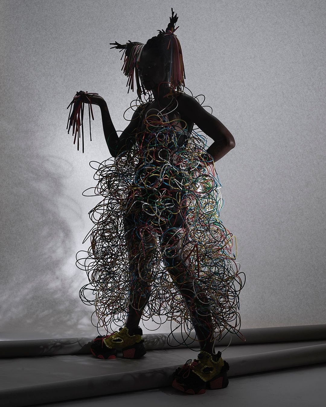 A person stands in a studio wearing a dress made of colorful wires, with matching hair and shoes, against a graduated gray backdrop.