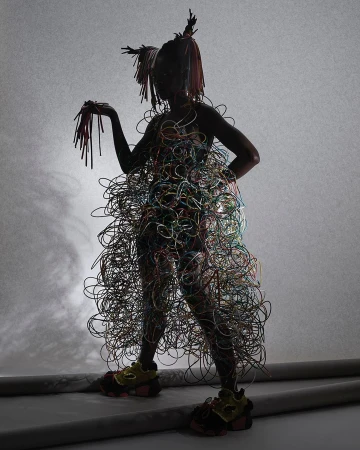 A person stands in a studio wearing a dress made of colorful wires, with matching hair and shoes, against a graduated gray backdrop.