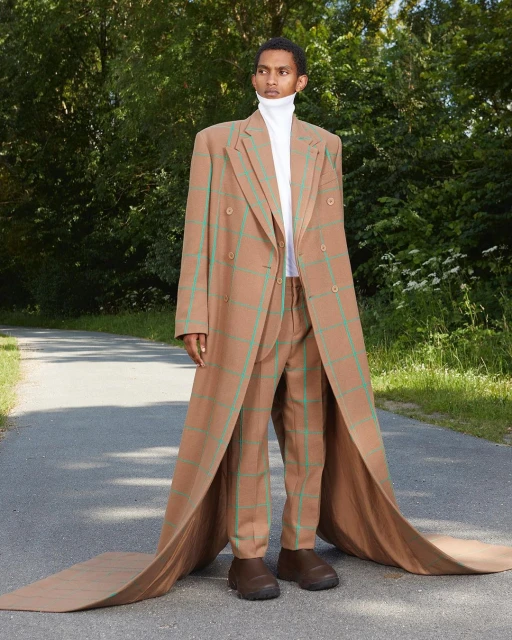 A man in a tan, plaid suit walks on a path surrounded by green trees. The oversized suit has a long train.
