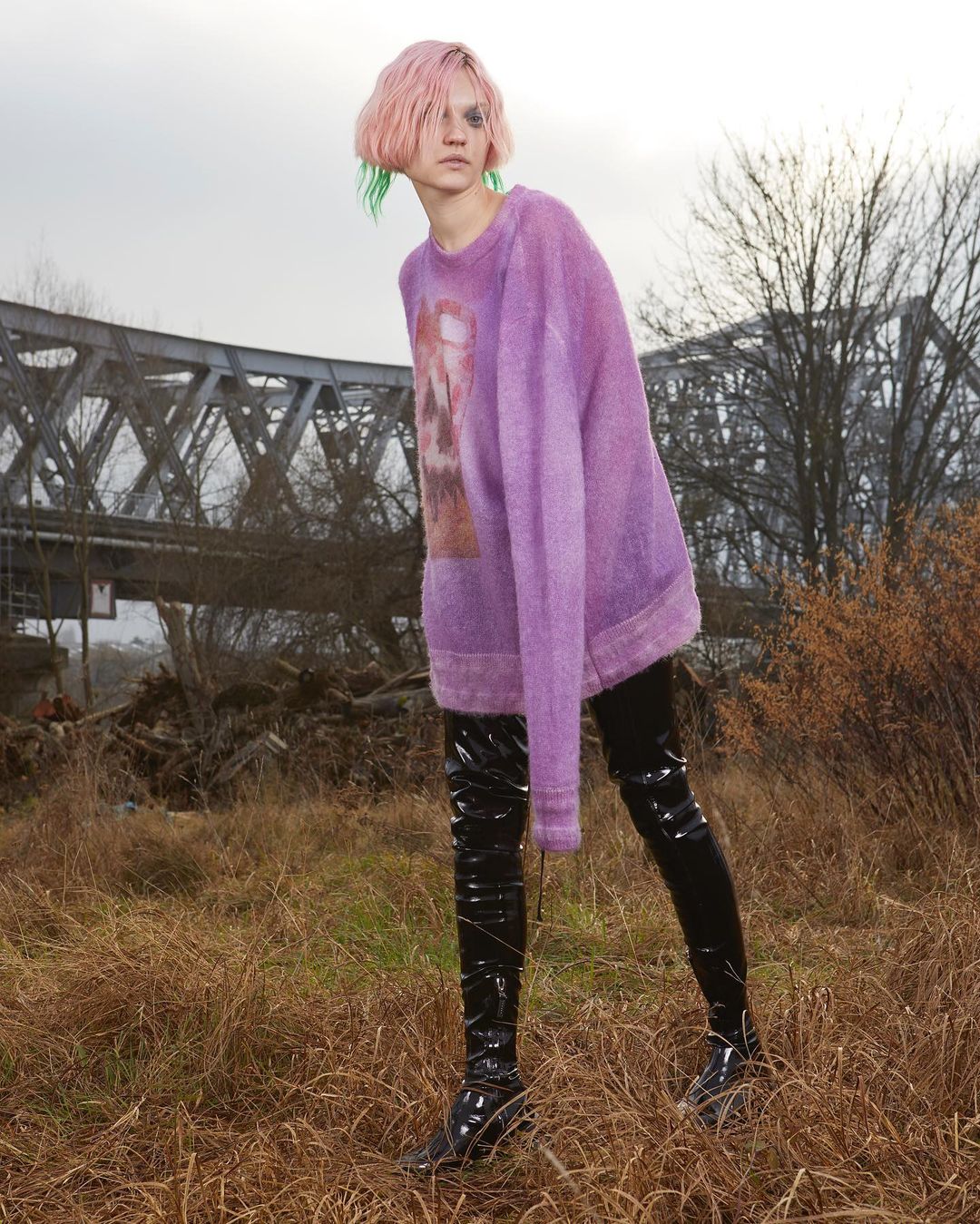 A young adult female with pink hair stands in a grassy field wearing a purple sweater, black shiny boots, and black leggings, posing for a fashion editorial.