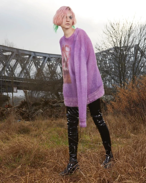 A young adult female with pink hair stands in a grassy field wearing a purple sweater, black shiny boots, and black leggings, posing for a fashion editorial.