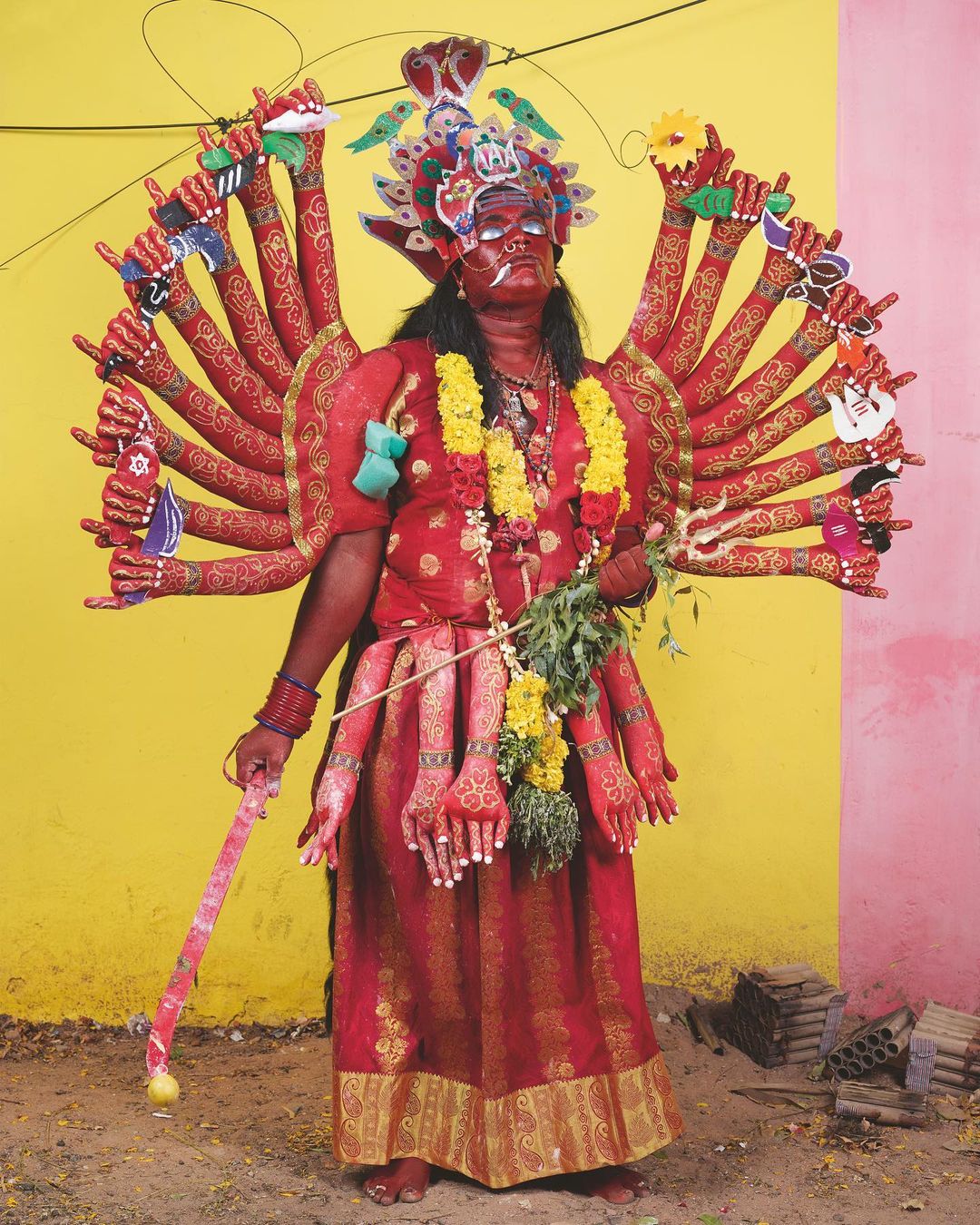 Adult in vibrant red Indian Kali costume with headdress and multiple arms holding props standing outdoors against a yellow and pink wall.