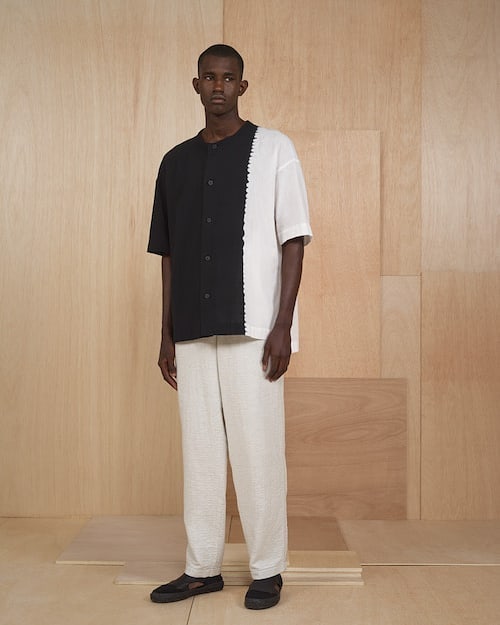Studio shot of a Black male model wearing a split black and white shirt and cream trousers, posing against a neutral plywood backdrop.