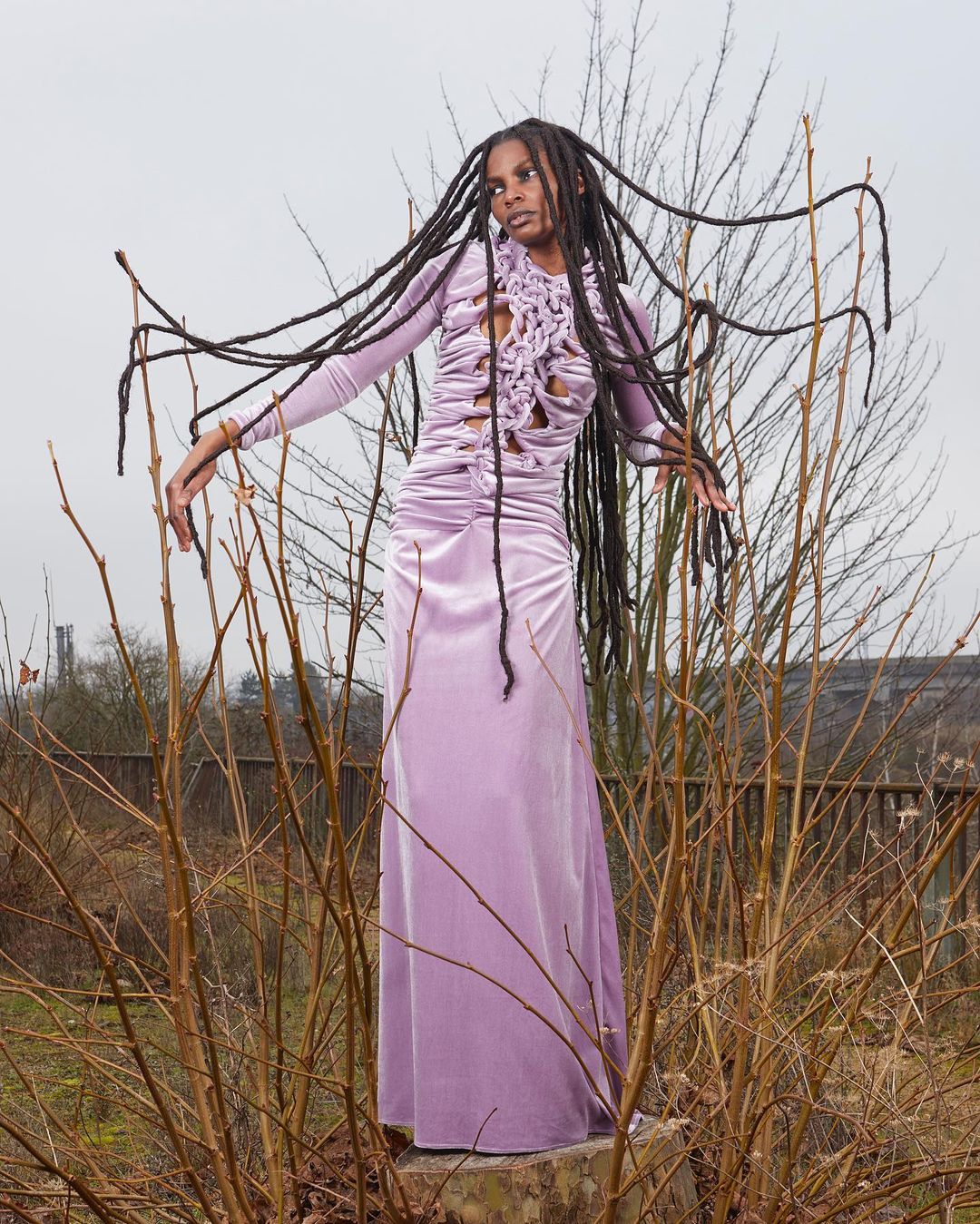 Full-length shot of a black female model in a lilac dress with dreadlocks, posing outdoors on a concrete block among bare branches.