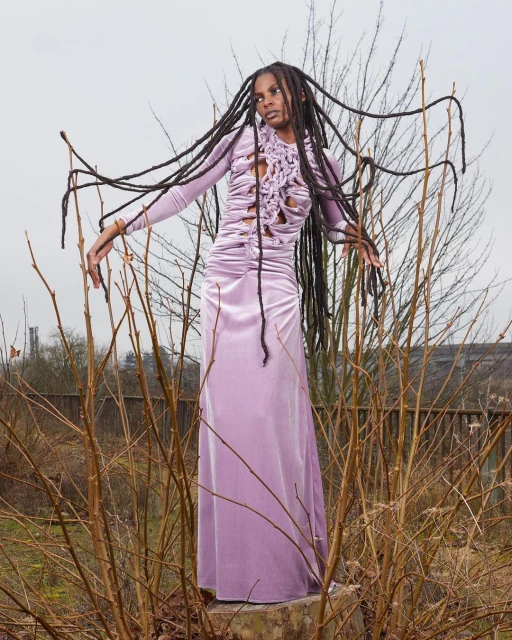 Full-length shot of a black female model in a lilac dress with dreadlocks, posing outdoors on a concrete block among bare branches.