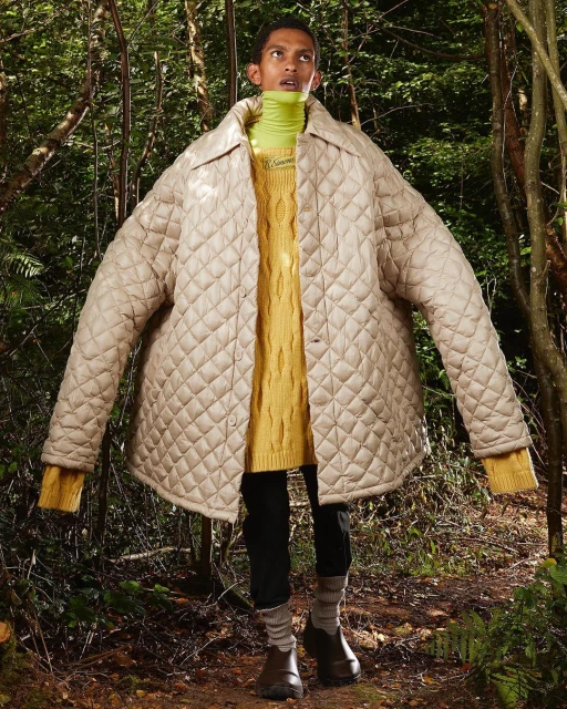 Full-shot of a person in the woods wearing a quilted jacket over a yellow sweater, black pants and brown shoes standing on foliage.