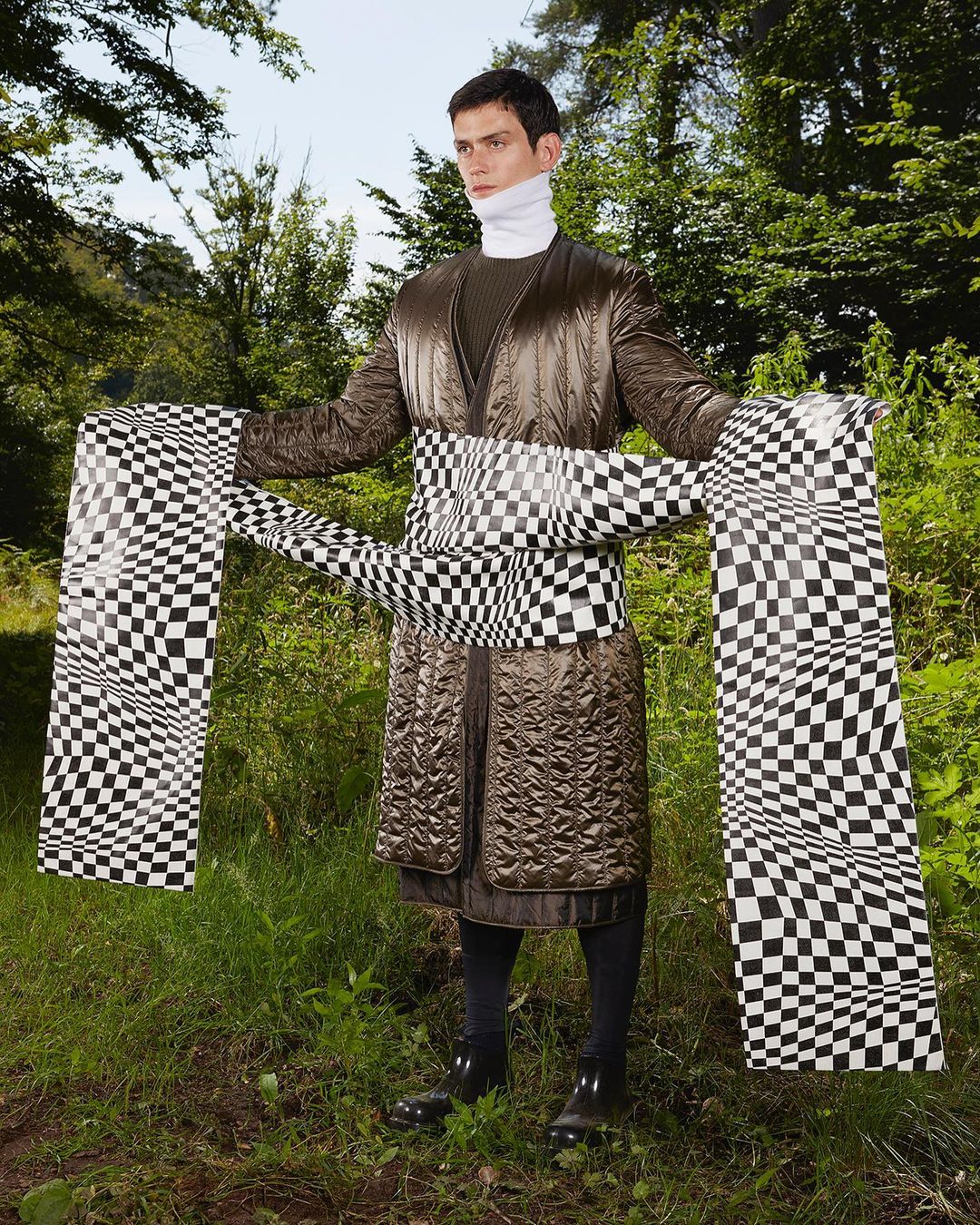 Adult male in brown quilted coat and checkered sash stands in a forest setting surrounded by green foliage and tall trees.