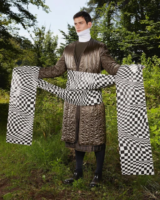Adult male in brown quilted coat and checkered sash stands in a forest setting surrounded by green foliage and tall trees.