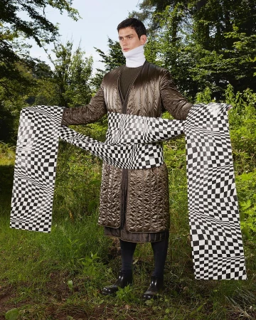 Adult male in brown quilted coat and checkered sash stands in a forest setting surrounded by green foliage and tall trees.