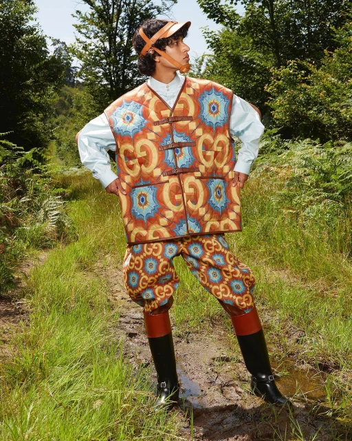Young adult posing in a patterned vest and pants, accessorized with a visor and boots, set against a lush, green forest backdrop.