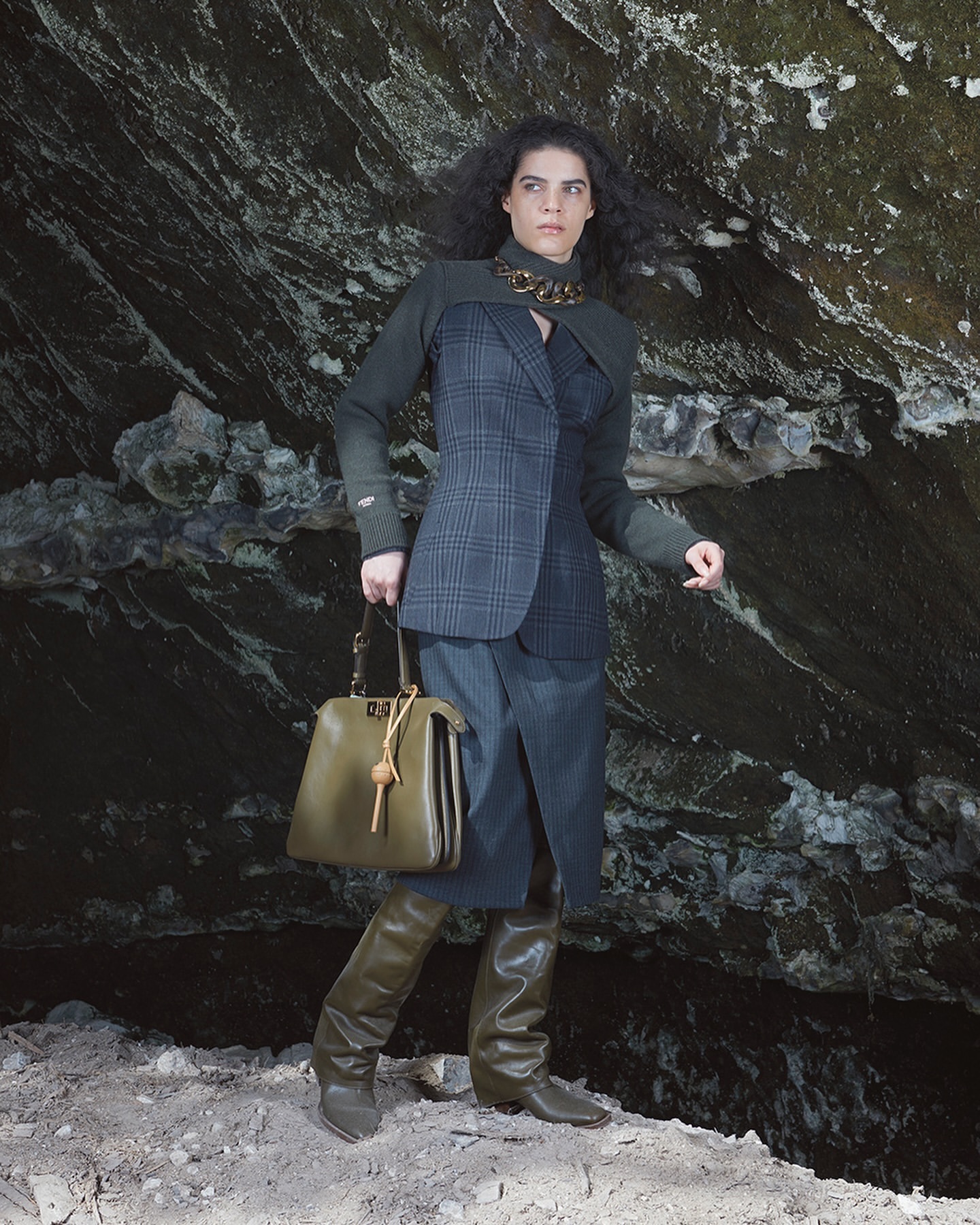 Adult female in a grey and green jacket with plaid blazer stands in front of a dark rock wall while holding a handbag.