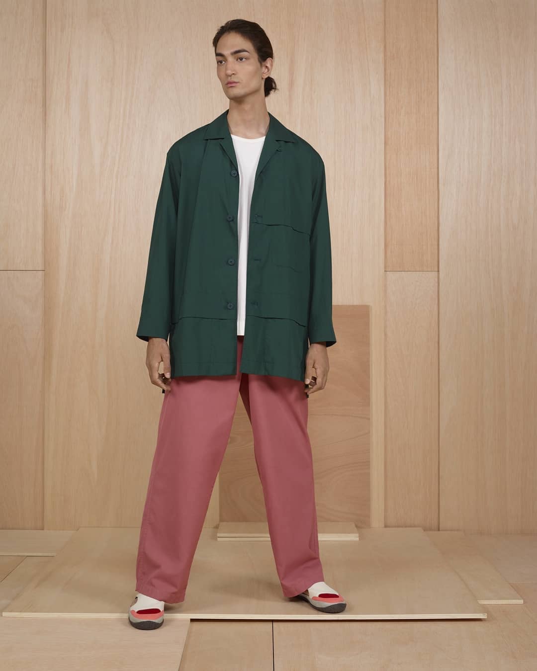 A person in a green jacket and mauve pants stands against a light wood paneled backdrop in a fashion or style editorial shot.