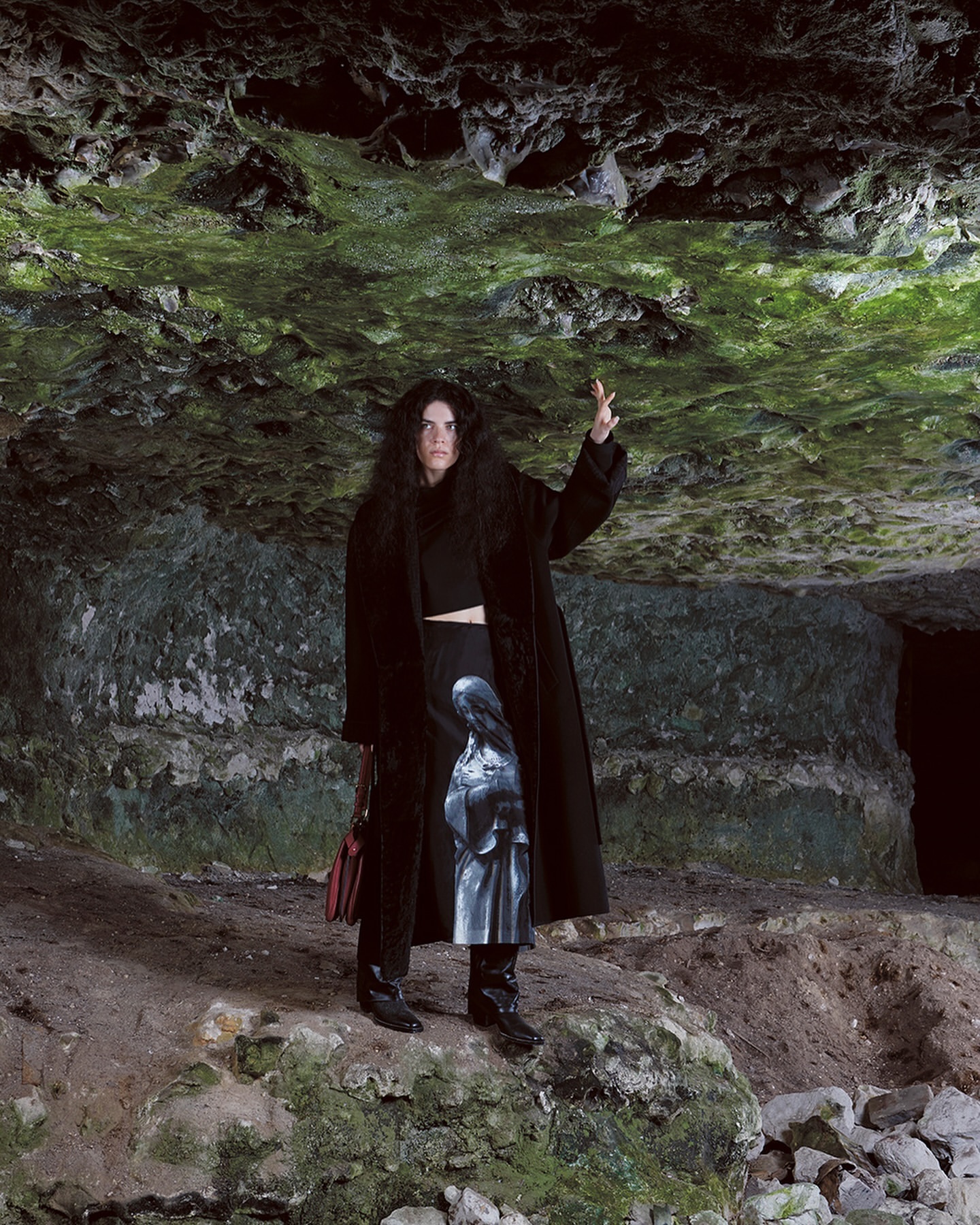 A woman in a black coat poses in a dimly lit cave with textured rock walls, creating a fashion editorial shot.
