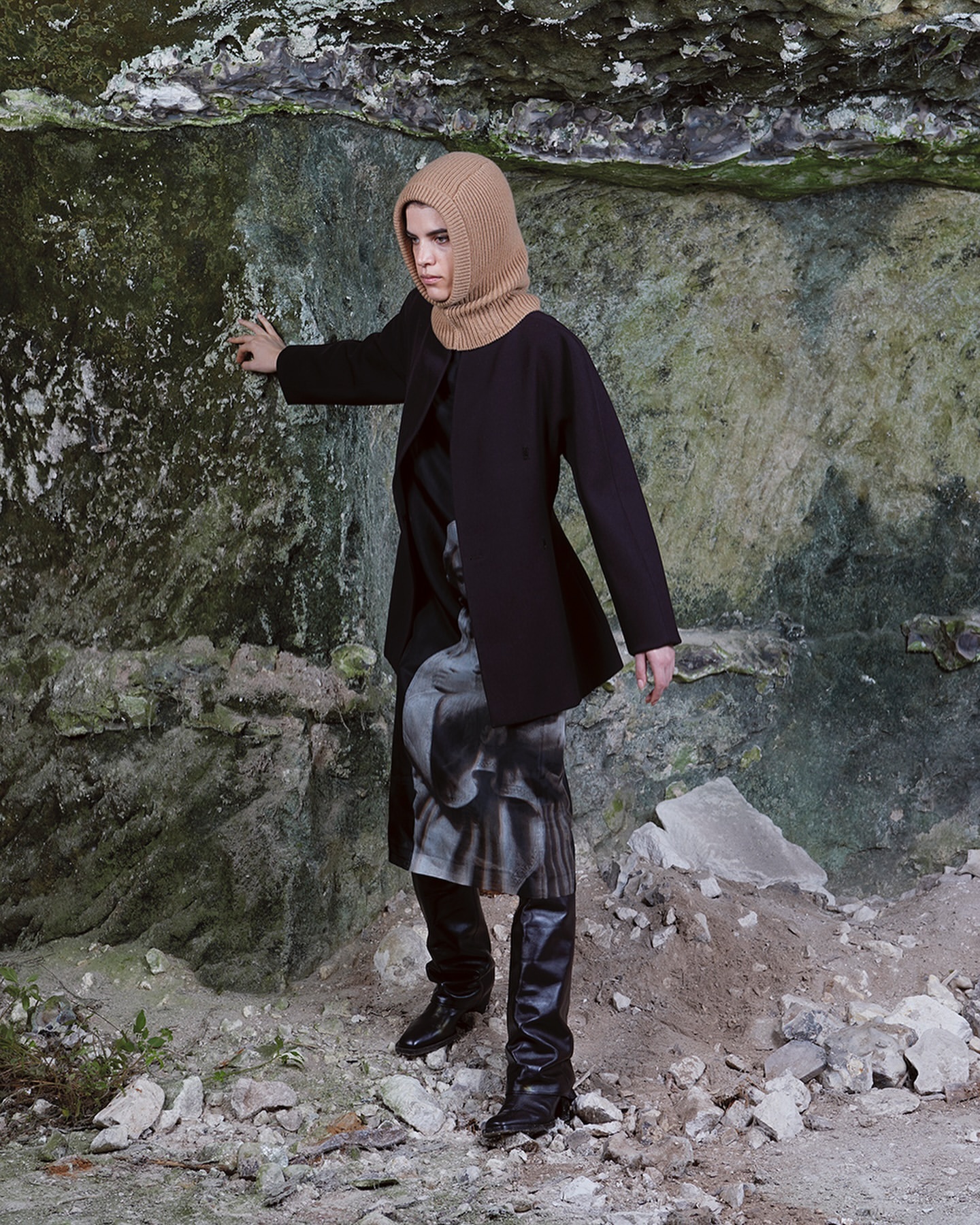 Model wearing a tan knit balaclava, black coat, and tall boots standing against a rough stone rock face in Paris.