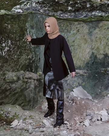 Model wearing a tan knit balaclava, black coat, and tall boots standing against a rough stone rock face in Paris.