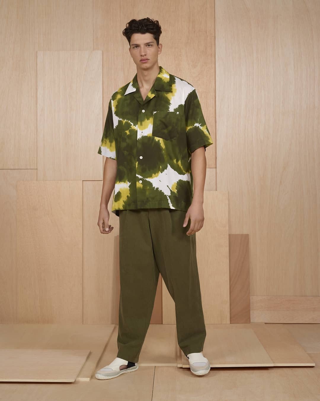 Male model in a relaxed pose, wearing an olive green and white tie-dye style short-sleeved shirt and olive green pants, on a wood panel background.