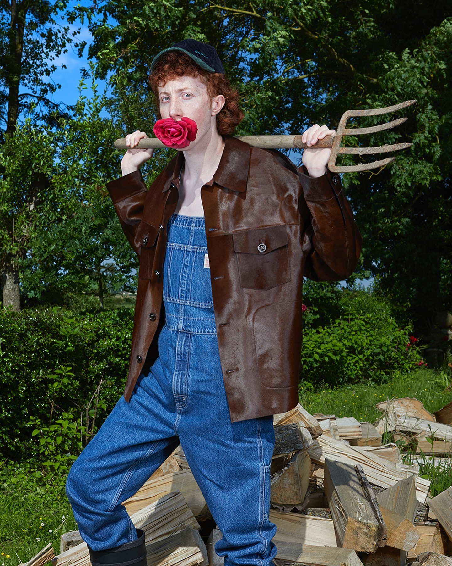 Person in overalls, wearing a leather jacket and cap, poses outdoors with a rose in their mouth and a pitchfork over their shoulders.