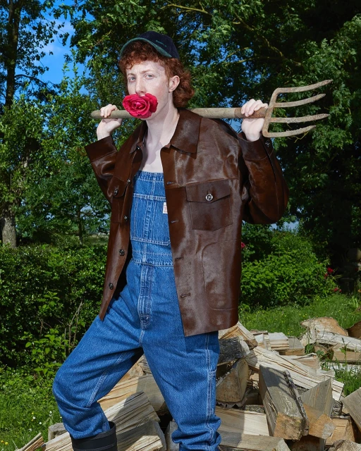Person in overalls, wearing a leather jacket and cap, poses outdoors with a rose in their mouth and a pitchfork over their shoulders.