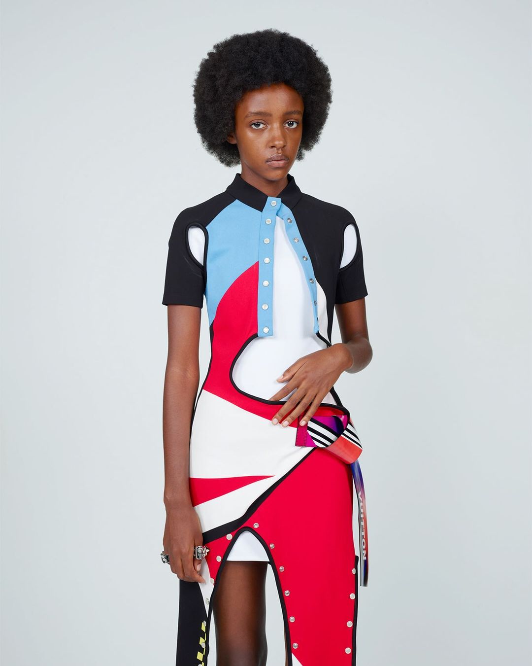 Adult female fashion model with afro poses in a studio wearing a color-blocked dress with geometric patterns and silver hardware accents.