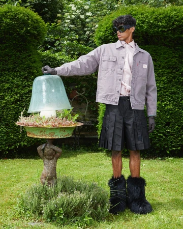 Man in a garden wearing a pale purple jacket and black kilt standing next to a lamp on a cherub stand with green foliage.