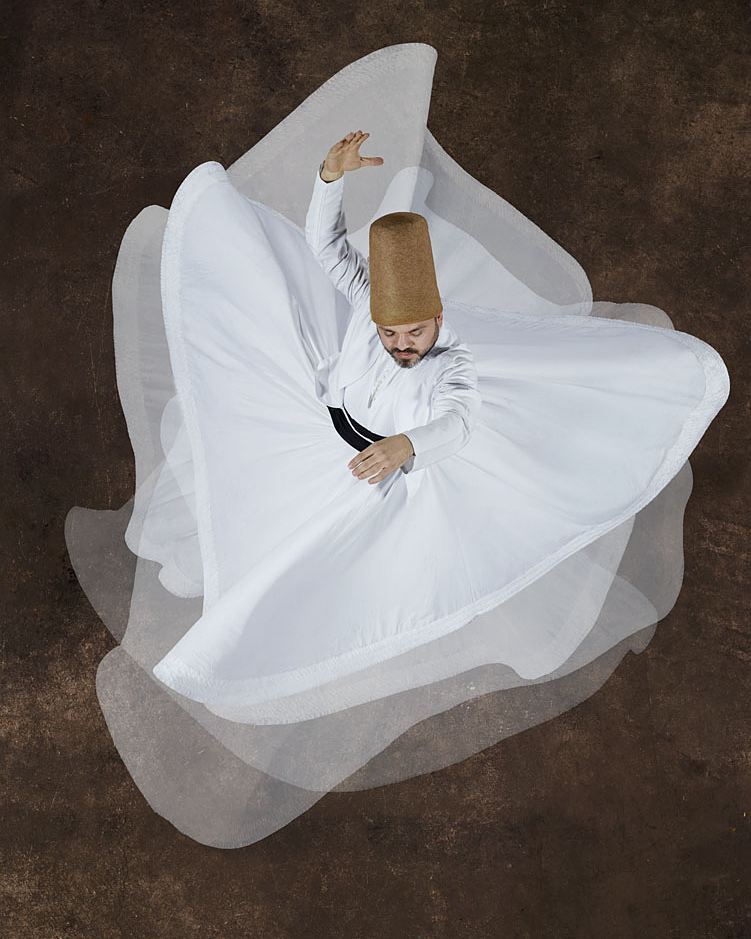 Man wearing a conical hat and white robe spins with arms outstretched against a mottled brown background in a top-down medium shot.