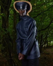Black man wearing dark leather jacket and matching cap with tan trim in a dense forest setting