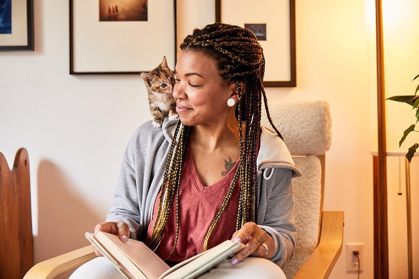 Woman in chair reading a book with a kitten perched on her shoulder in an indoor setting with artwork and a floor lamp.