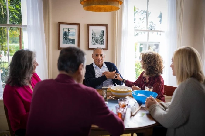 Five people are gathered around a table in a brightly lit room with large windows, sharing snacks and drinks in a casual setting.