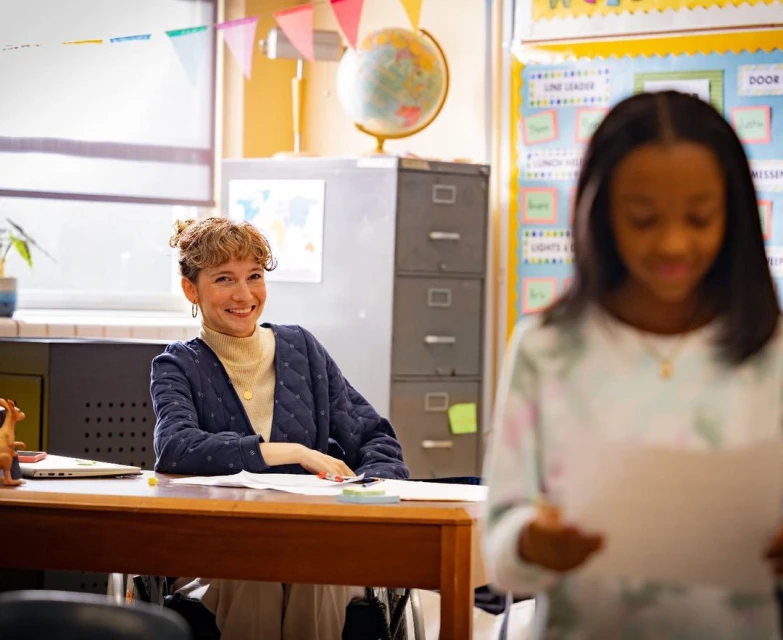 Teacher smiling at an elementary school student presenting a paper in a classroom with desks, flags, globe, and filing cabinet.