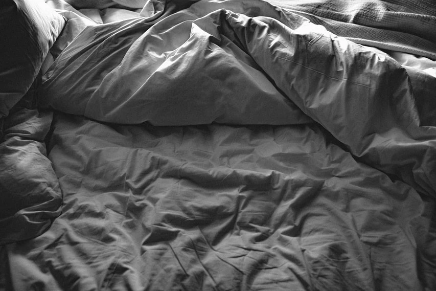 Monochrome image displaying a rumpled bed with disarranged sheets and pillows, suggesting a recently slept-in or unmade bed, indoors.