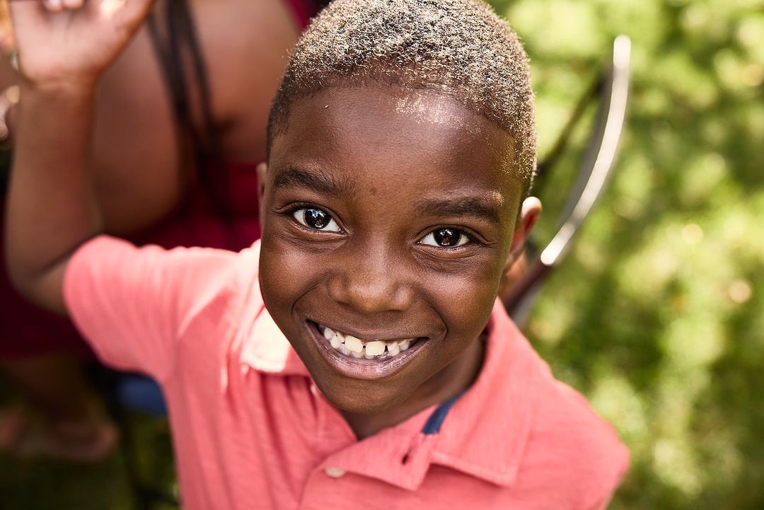 Close-up portrait of a smiling Black child in a coral shirt, looking directly at the camera in an outdoor, natural setting.