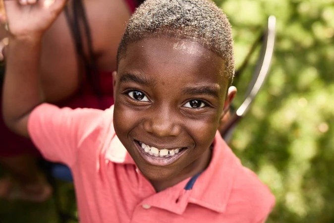 Close-up portrait of a smiling Black child in a coral shirt, looking directly at the camera in an outdoor, natural setting.