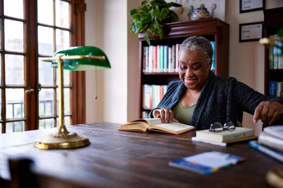 Senior African-American woman reading book at wooden table with glasses, lamp, and bookshelfs in background, indoor setting.