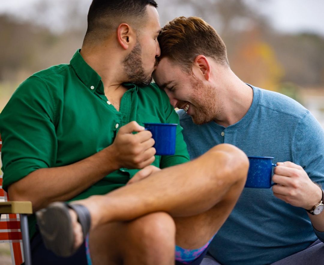 Two men outdoors, one kissing the other's forehead, each holding a blue mug in a candid and intimate setting.