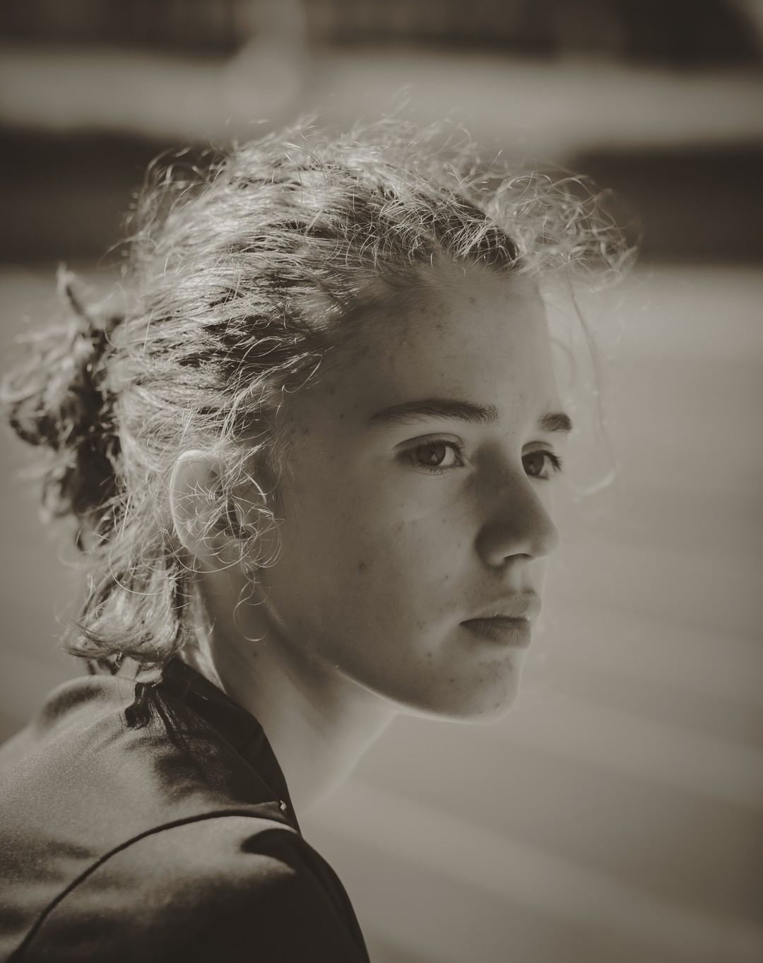 Monochrome portrait of a young fair-skinned person with curly hair, looking off-camera, in a blurry outdoor environment.