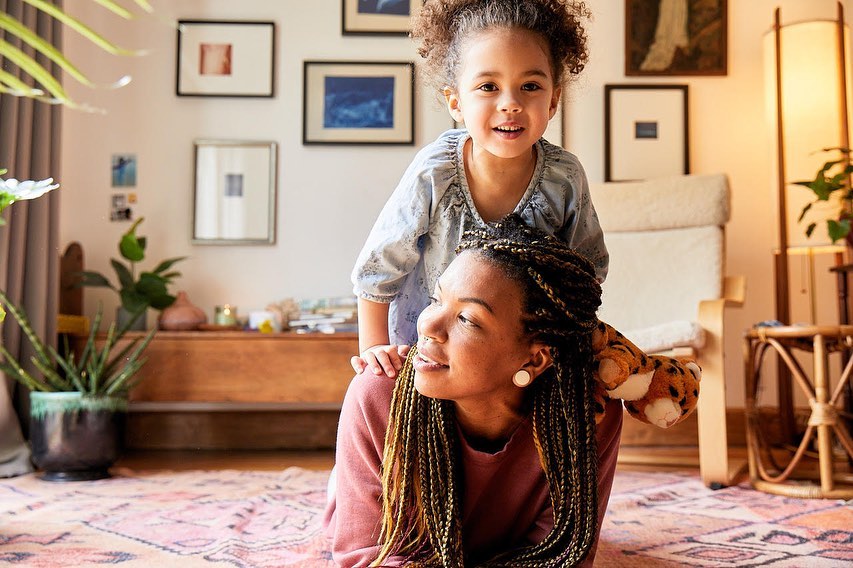 Smiling child sits on the back of an adult woman in an indoor living room setting with plants and a patterned rug in the frame.
