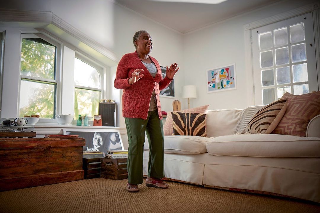 Senior woman dances with joy at home in a red cardigan and green pants. The living room has a couch and large window with natural light.