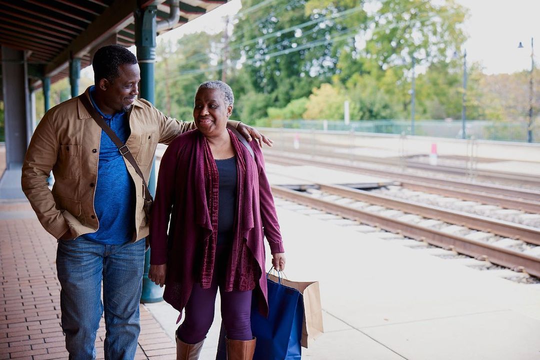 Two people, an adult man and an elderly woman, are standing on a train platform. The man has his arm around the woman's shoulders.