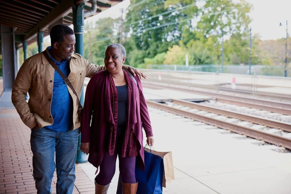 Two people, an adult man and an elderly woman, are standing on a train platform. The man has his arm around the woman's shoulders.