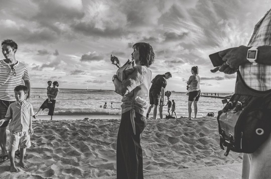 Black and white photo of people on a sandy beach with waves, clouds, and distant figures creating a dynamic scene.