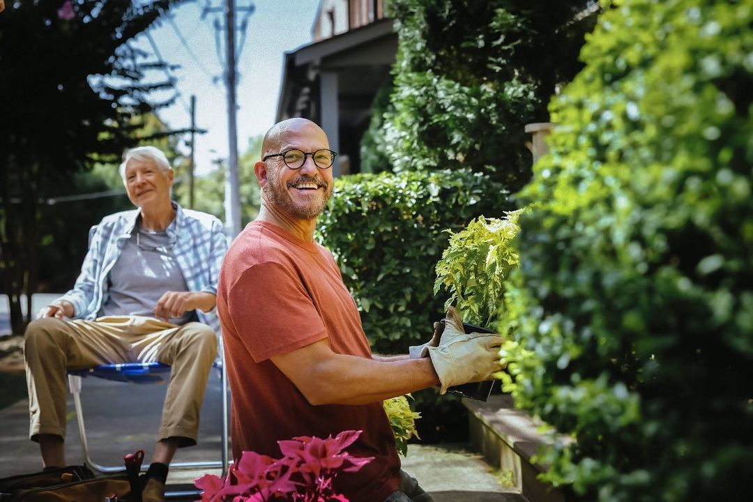 Two men outdoors: one bald man in an orange shirt tending plants, smiling; one elderly man sits in background.