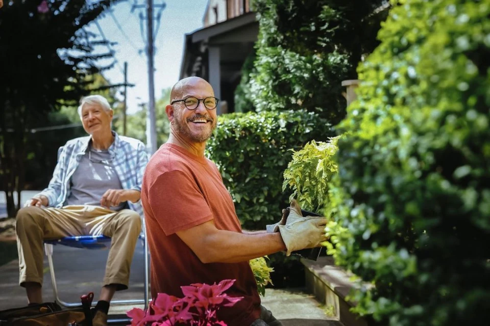 Two men outdoors: one bald man in an orange shirt tending plants, smiling; one elderly man sits in background.