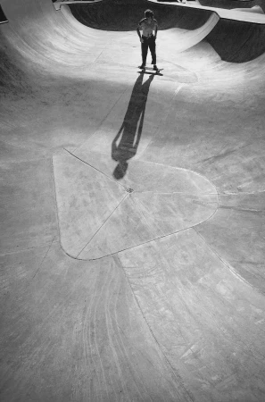 Monochrome high-angle view of a skateboarder standing in a concrete bowl casting a long shadow towards the camera.