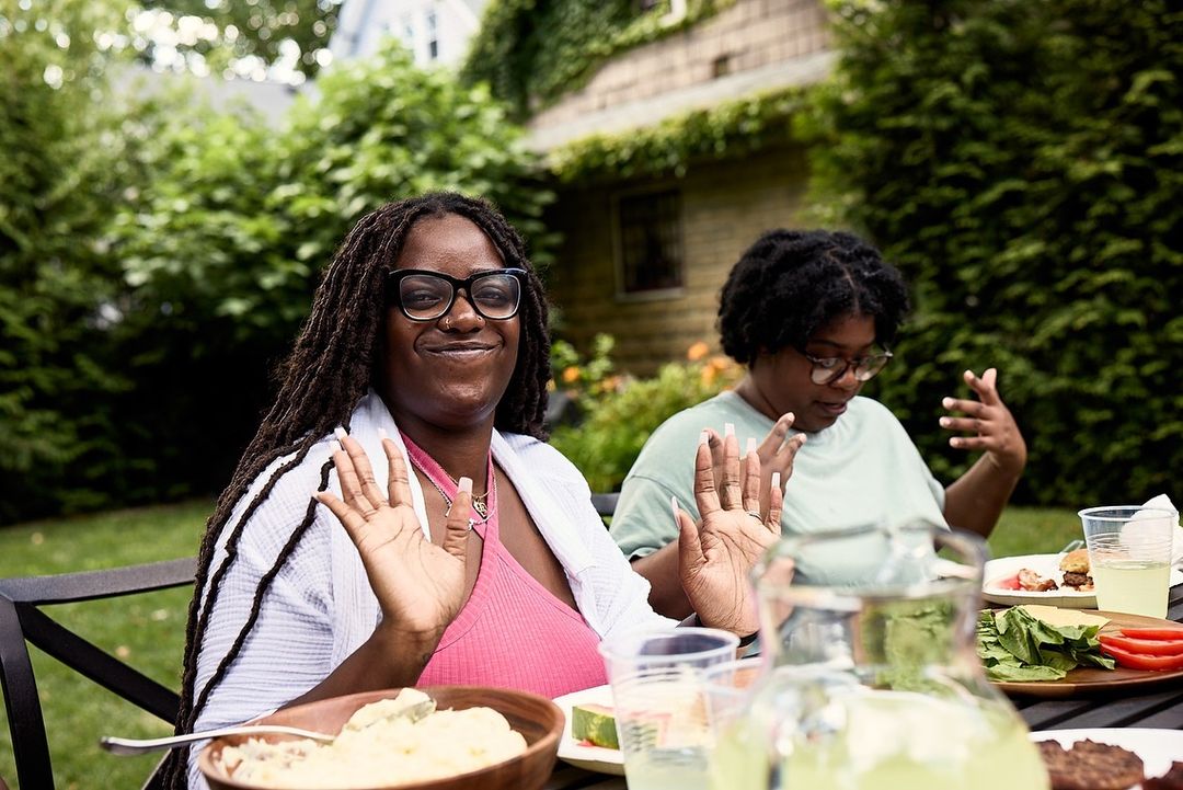 Two women smiling outdoors at a picnic table. One woman is in focus with glasses and long hair, the other is out of focus.