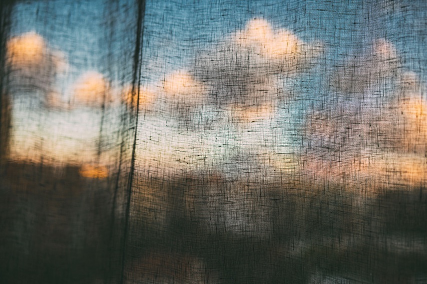 Abstract view through a sheer curtain reveals a colorful sky with clouds. Texture and light interplay in an atmospheric scene.