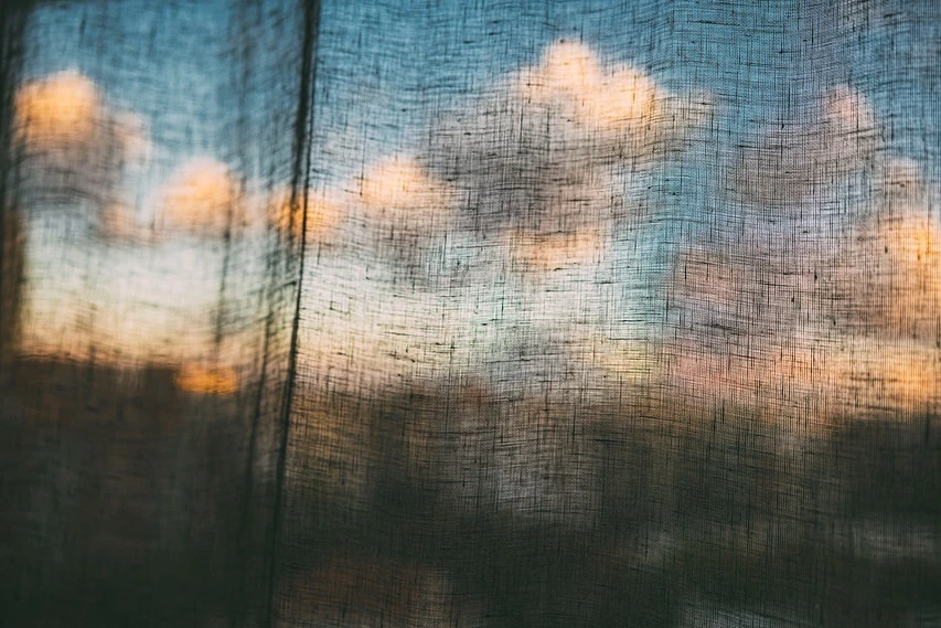 Abstract view through a sheer curtain reveals a colorful sky with clouds. Texture and light interplay in an atmospheric scene.