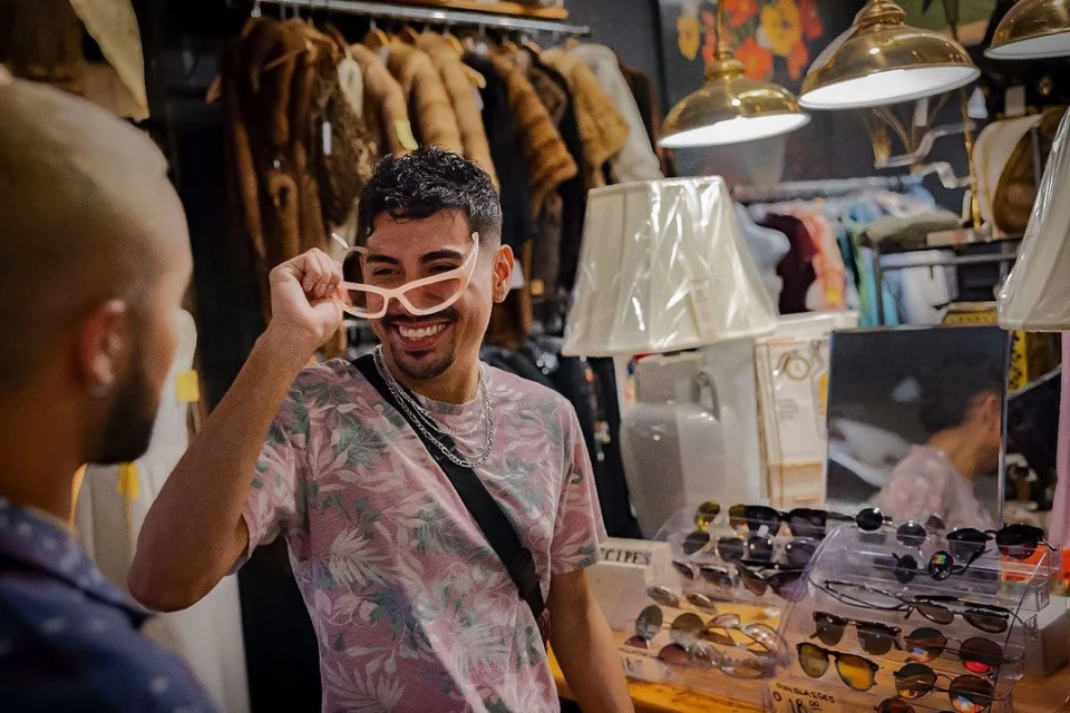 Two people in a vintage clothing shop. A man holds up sunglasses while smiling. Racks of clothing, lamps, and sunglasses displays surround them.